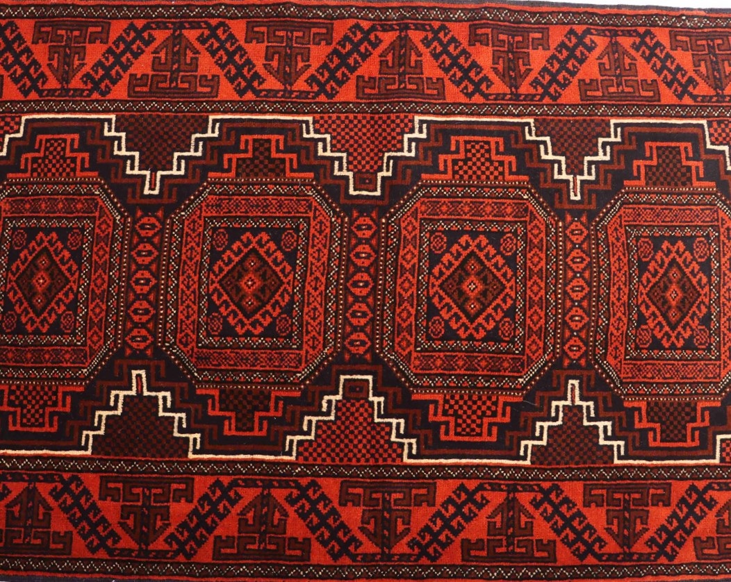 7 Compelling Reasons to Adorn Your Home with a Persian Rug 7 A close-up image of a handmade Balouchi rug featuring a symmetrical pattern with a vibrant color palette of red, blue, and cream. The intricate design includes diamond-shaped motifs and geometric patterns. The rug is placed on a hardwood floor with natural lighting casting shadows over its texture.