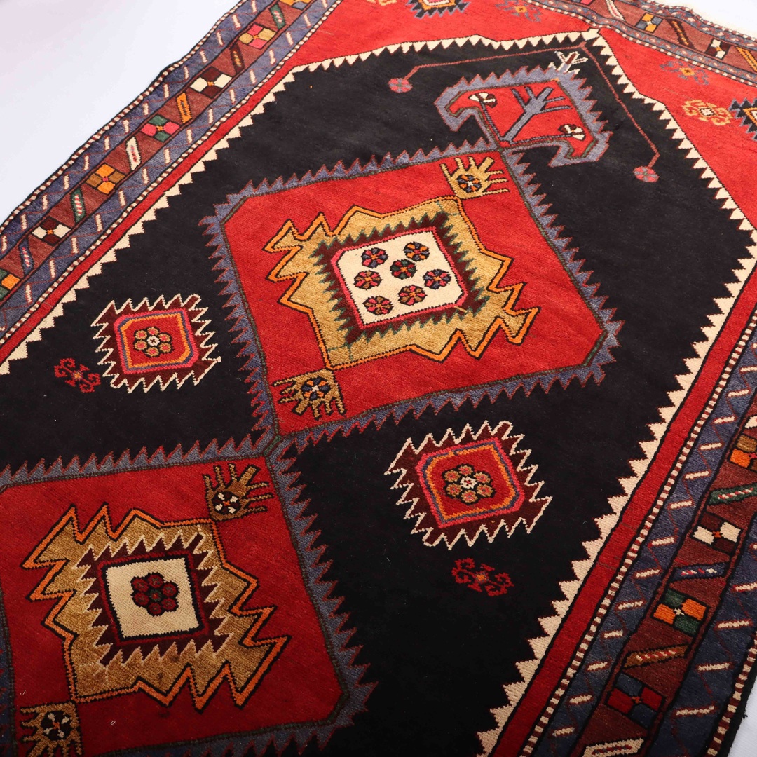 7 Compelling Reasons to Adorn Your Home with a Persian Rug 5 A close-up photo of a Persian rug with intricate geometric patterns and bright colors, lying on a wooden floor. The rug features a central diamond-shaped medallion in red, surrounded by smaller diamond-shaped motifs in blue, green, and beige. The edges of the rug are adorned with a series of thin borders in various colors and patterns.